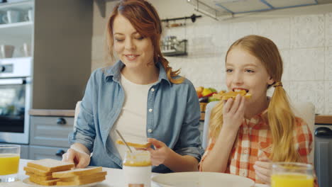 Portrait-shot-of-the-pretty-mother-doing-sandwiches-with-peanut-butter-and-her-lovely-daughter-eating-them-with-a-smile.-Indoor